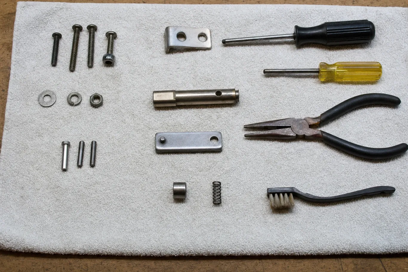 Machine screws, washers, roll pins, two stainless brackets and a small coil spring laid out on a grey towel beside nut drivers, long-nose pliers and a wire brush