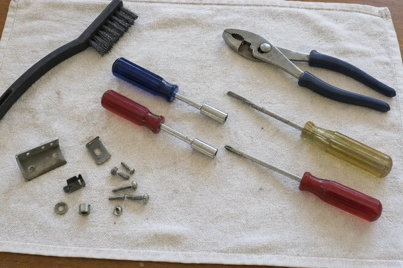 Two nut drivers with hex sockets fitted, slip-joint pliers, a wire brush and two screwdrivers on a cream towel beside small steel brackets, screws, washers and a threaded insert